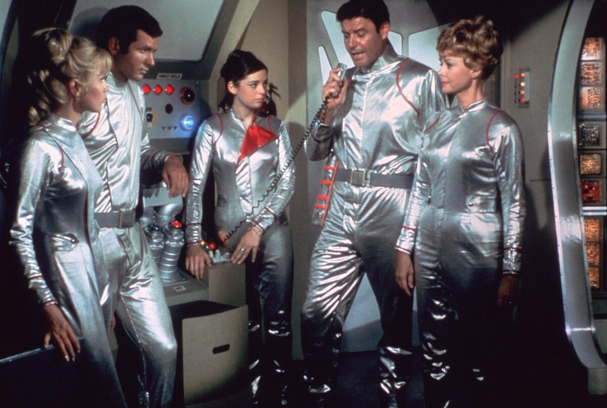 In a cast photo from "Lost in Space," five actors in silver spacesuits stand in a space ship.