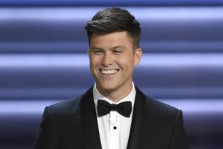 Colin Jost wearing a tuxedo and smiling against a blue blue background