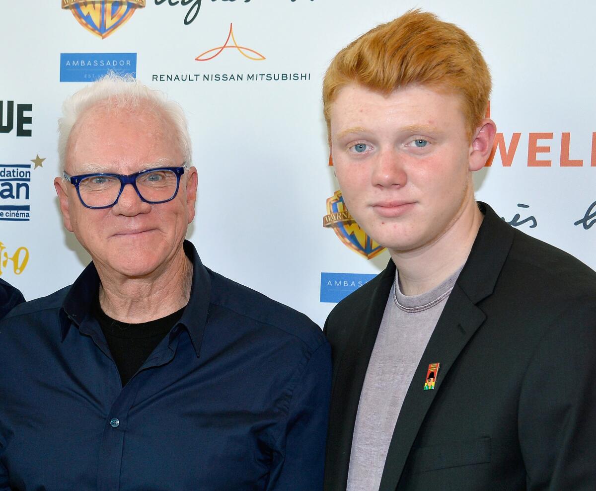 A white-haired man wearing glasses stands beside a young red-haired man wearing a suit jacket