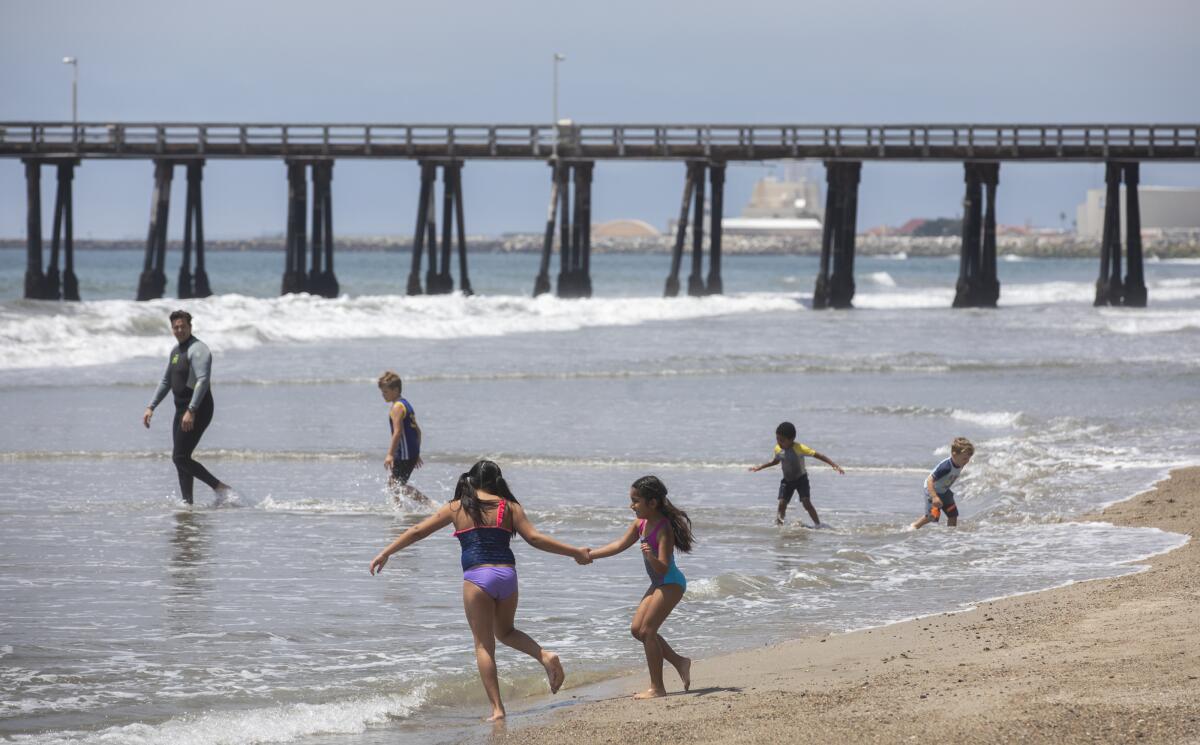 People play in the shallow water at a beach with a pier in the background