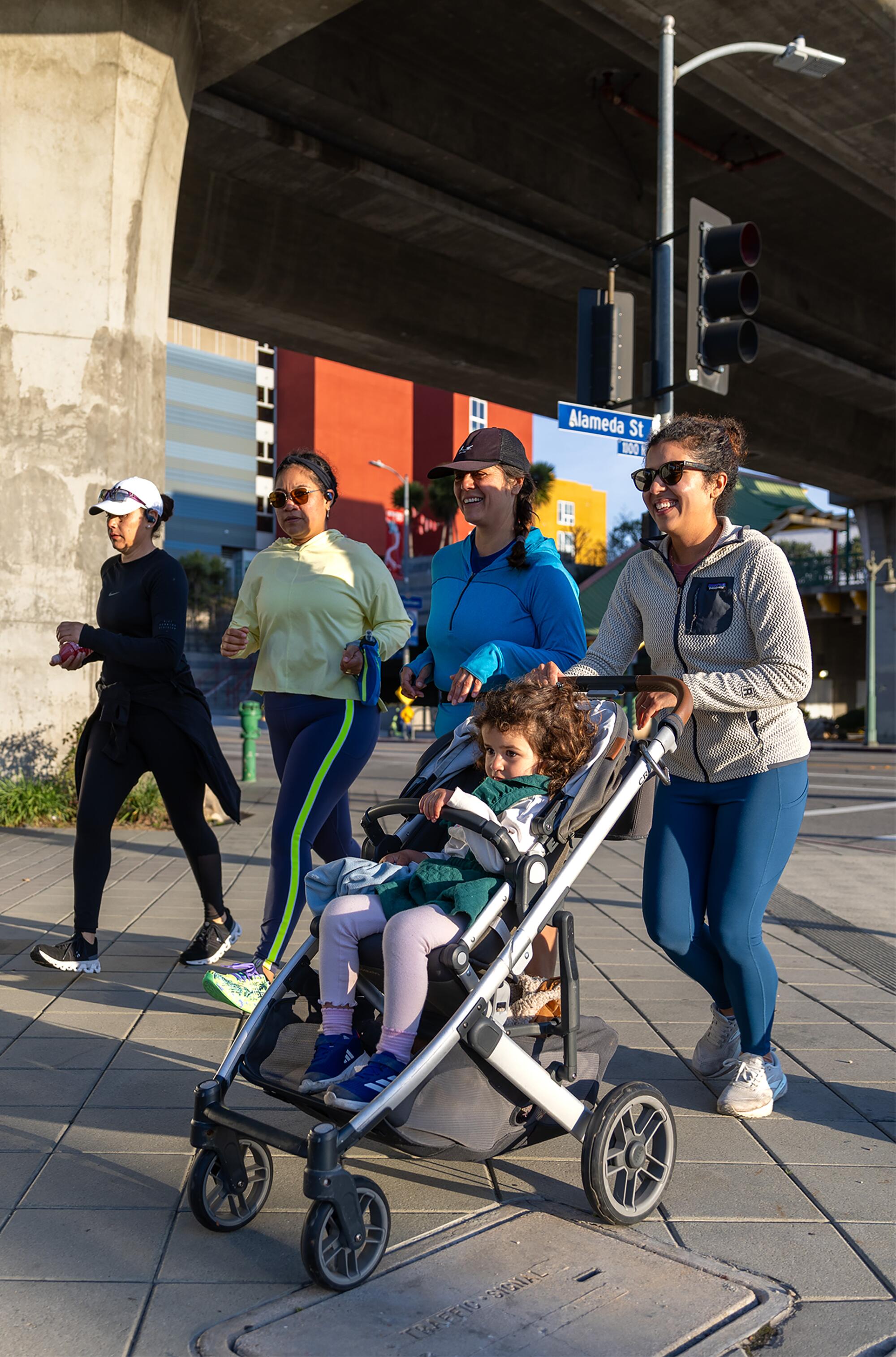 A woman pushes a child in a stroller as she and three other women walk or run outdoors.