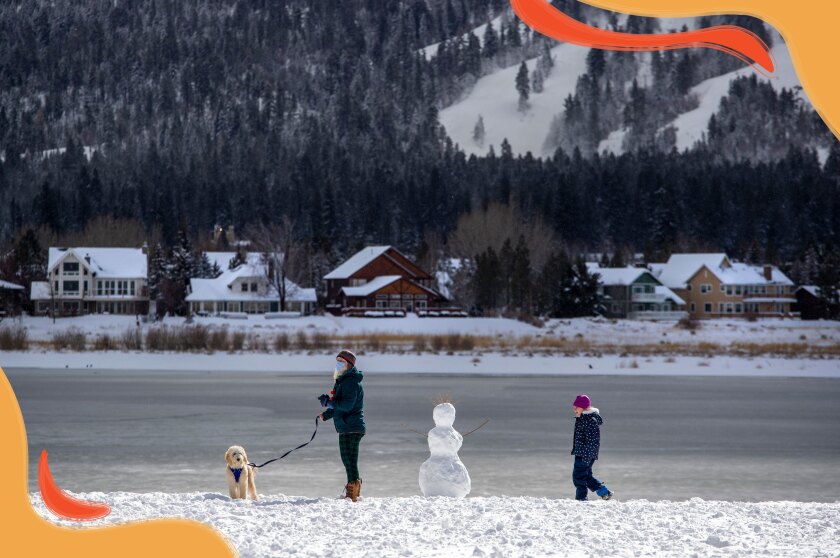A woman and a child stand on a snowy lakeshore with a dog and a snowman. Houses and evergreens are seen across the lake.