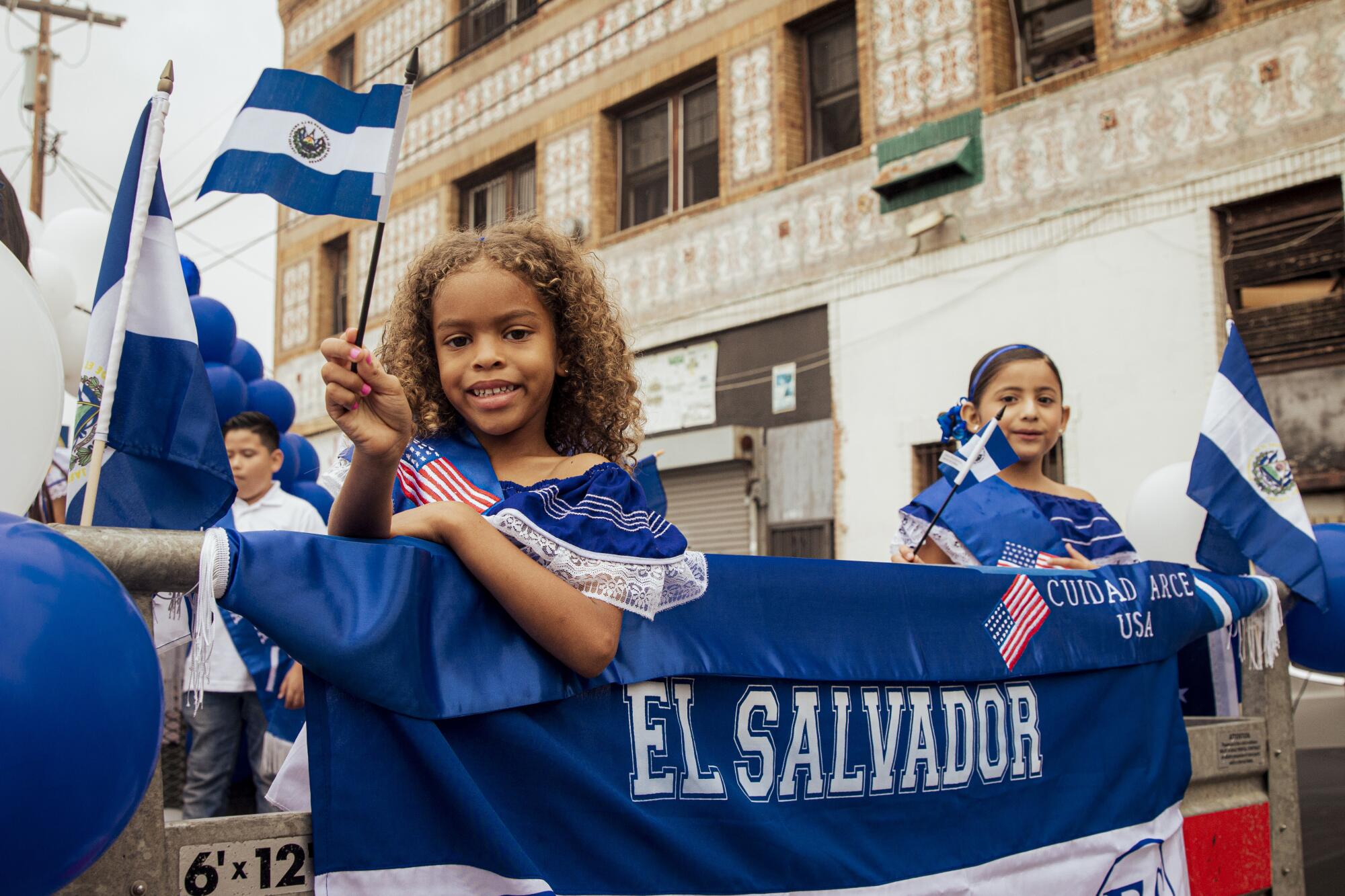 Two young girls stand on top of a float waving El Salvador flags