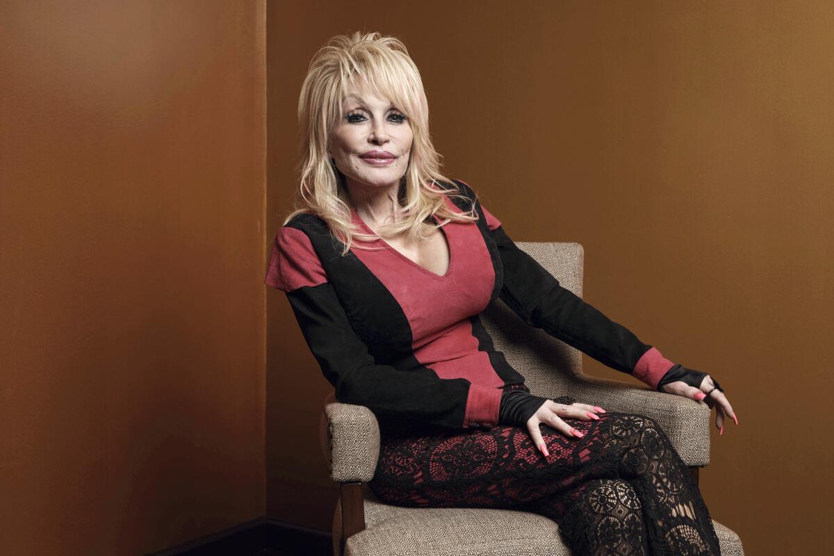Dolly Parton wears a red and black blouse with black fishnet leggings and teased blond hair. 