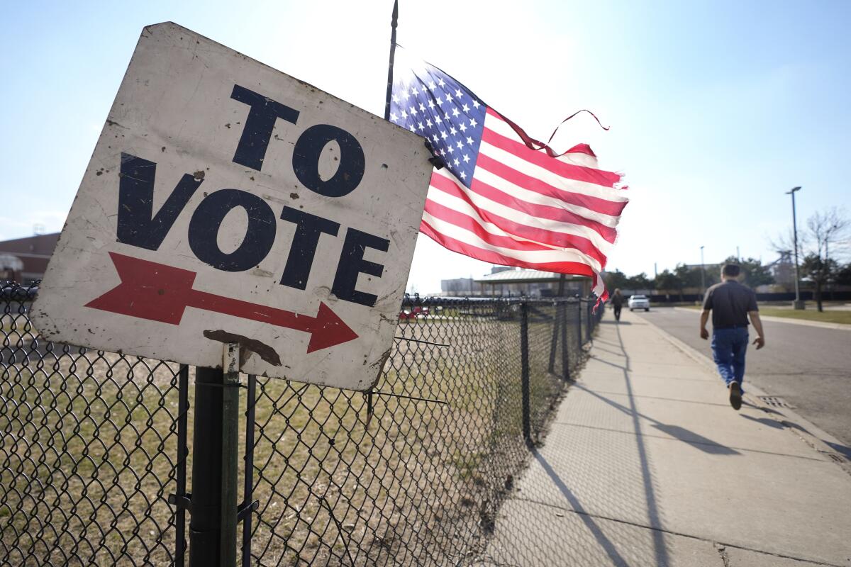 A voter walks on a sidewalk by a fence, a flag and a sign pointing the way to a primary election location. 