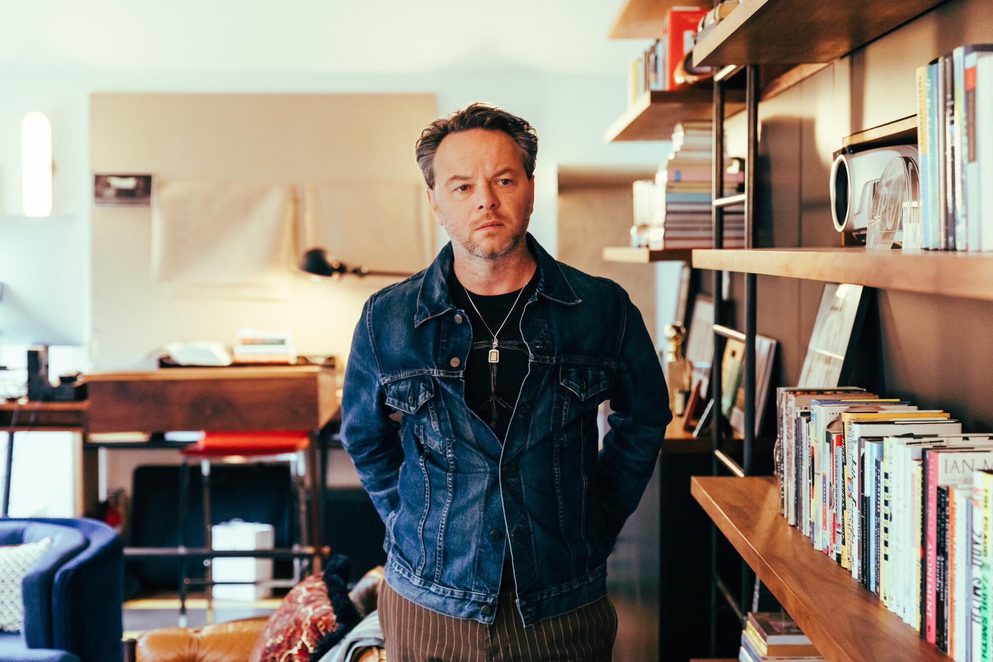 Noah Hawley in a denim jacket and black T-shirt stands next to bookshelves in his office.
