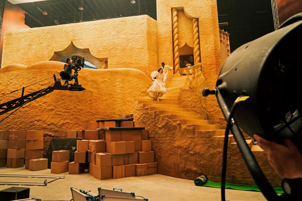 A film set shows two people running down gold-tinted stairs as a camera mounted on a crane takes the photo.