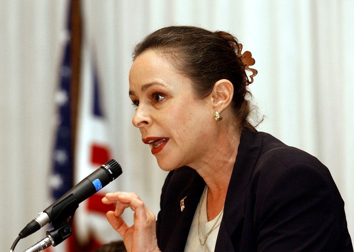 Alina Fernández, wearing a black jacket, her brown hair pinned in a clip at the back of her head, speaks into a mic