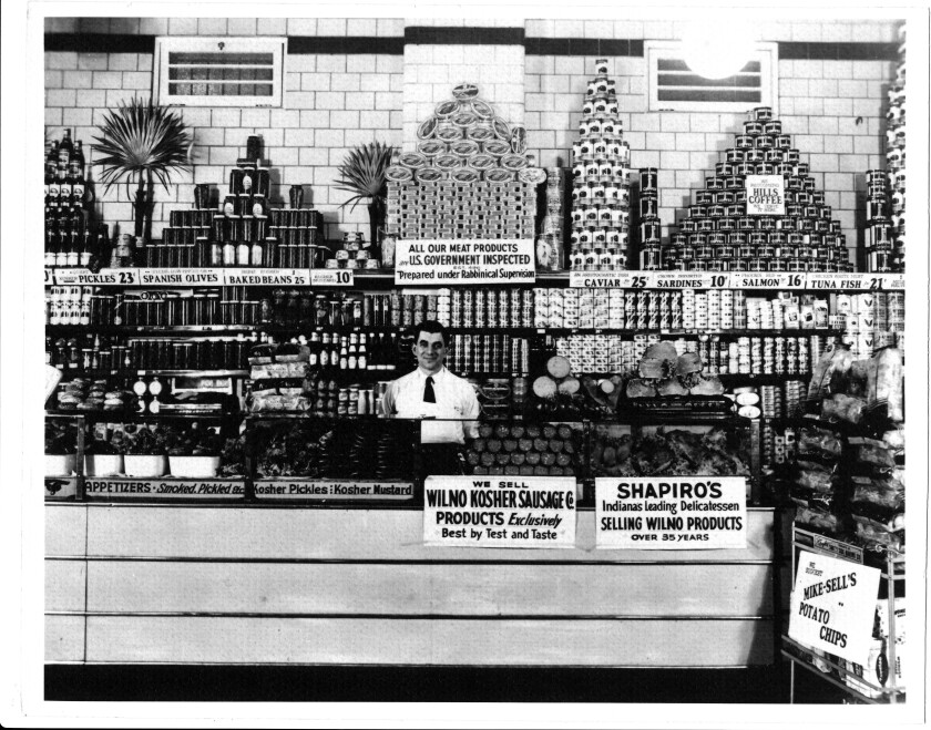 A man stands behind the counter of a delicatessen circa 1932.