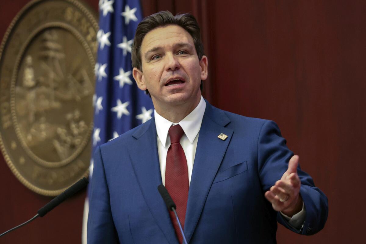 Ron DeSantis gestures while speaking into microphones, a large bronze state seal on the wall and an American flag behind him.