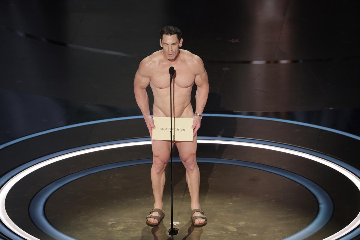 John Cena nude, wearing sandals and holding a big envelope in front of his groin