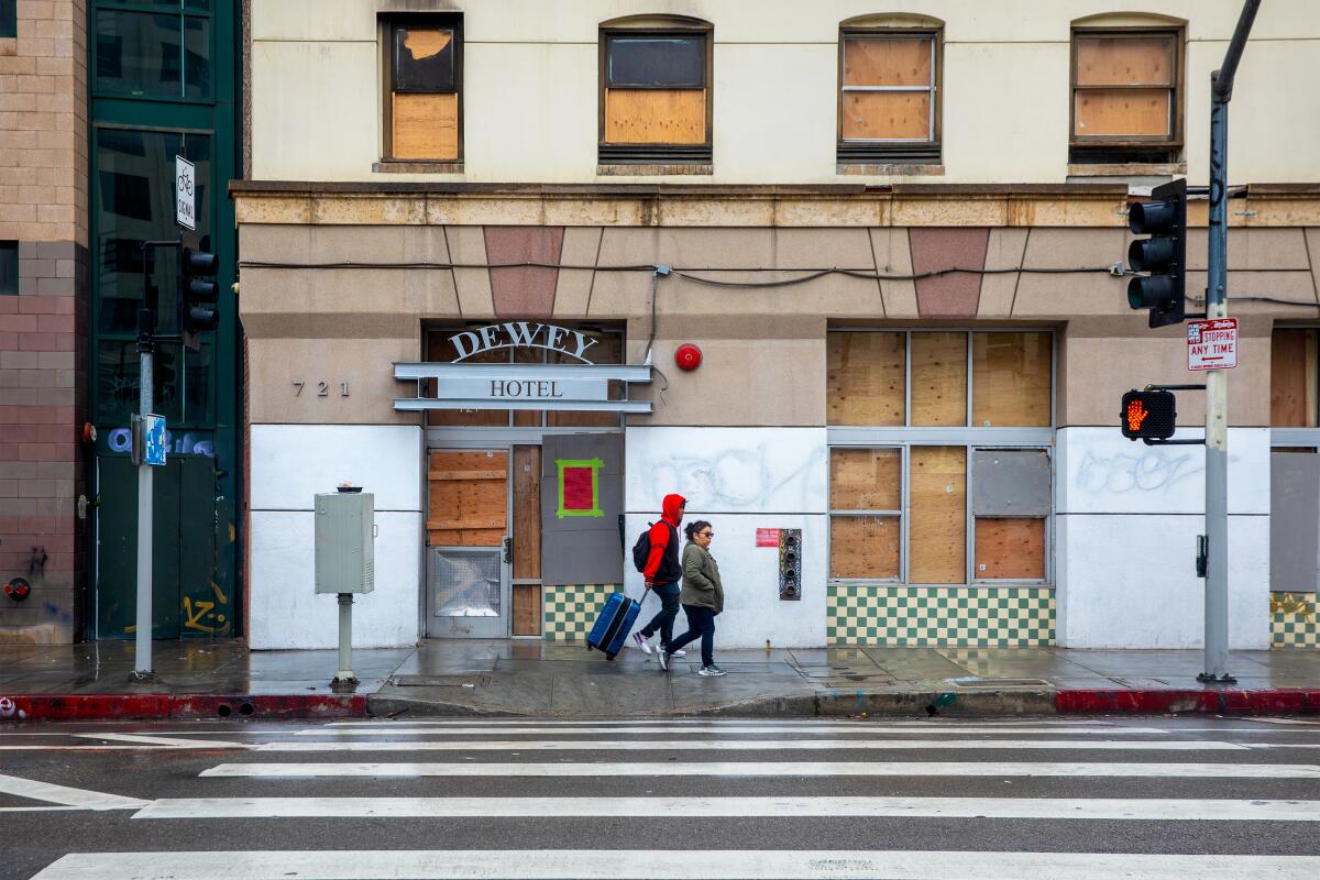 Two pedestrians pass a building with boarded-up windows and a sign reading "Dewey Hotel"
