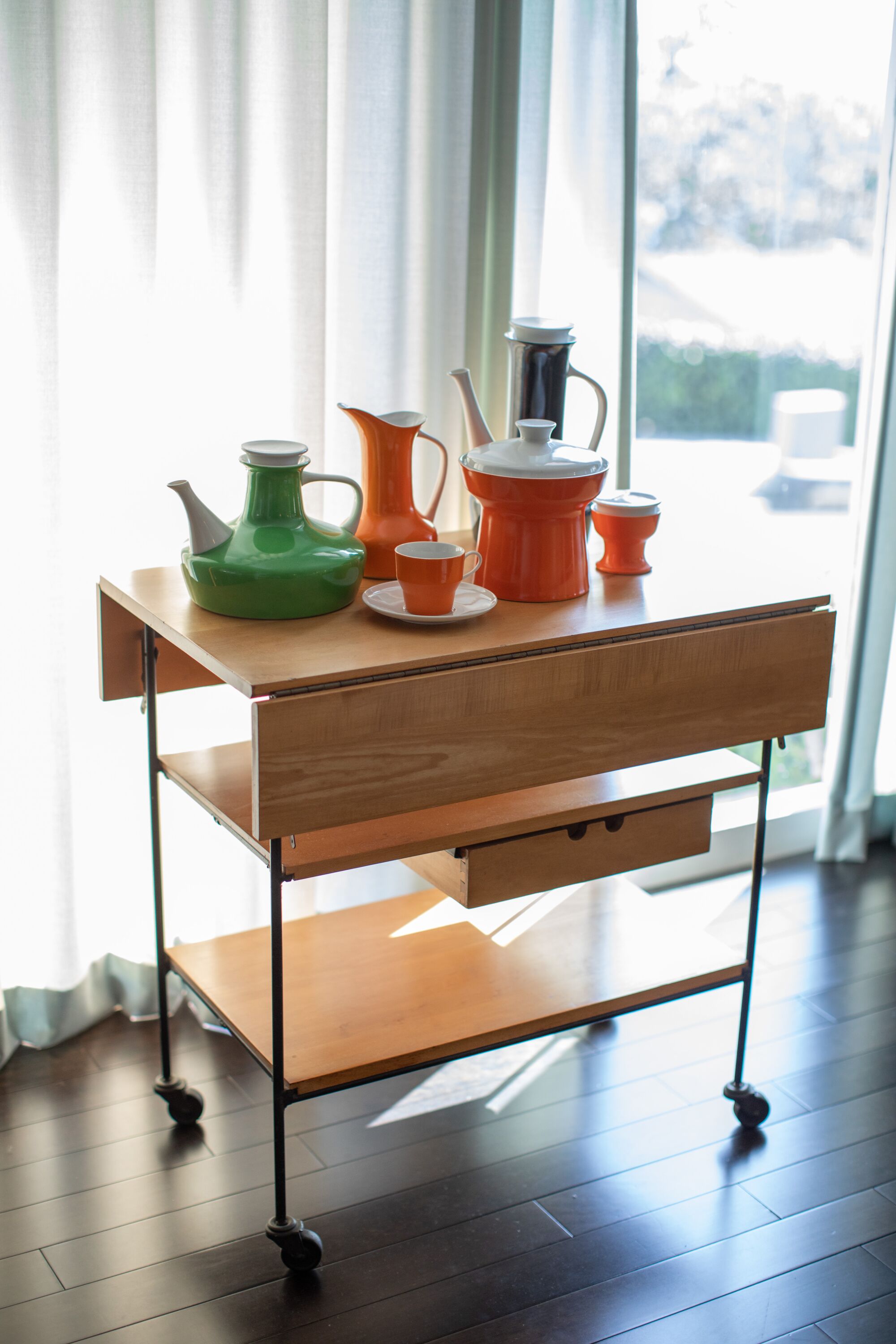 A Modernist tea wagon made from wood (resembling a bar cart) is shown bearing colorful ceramics in sunlight.