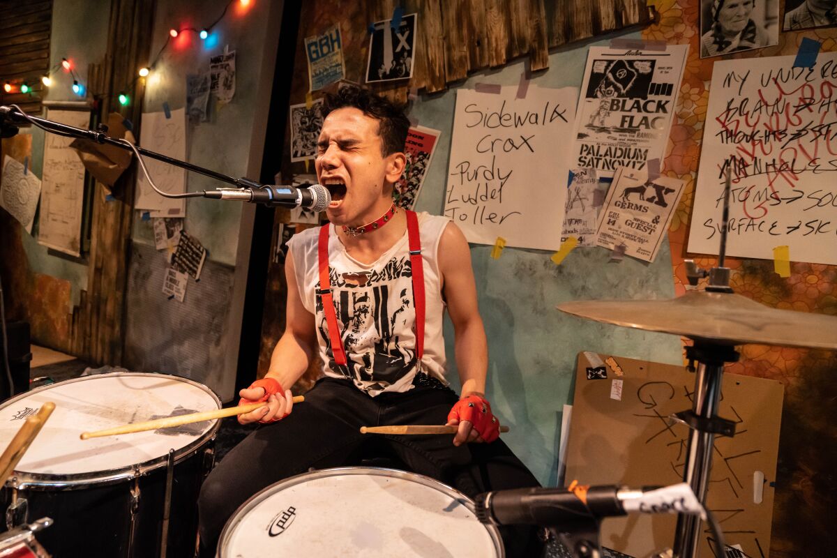 A man behind a drum set wearing red suspenders squints his eyes as he yells into a microphone.