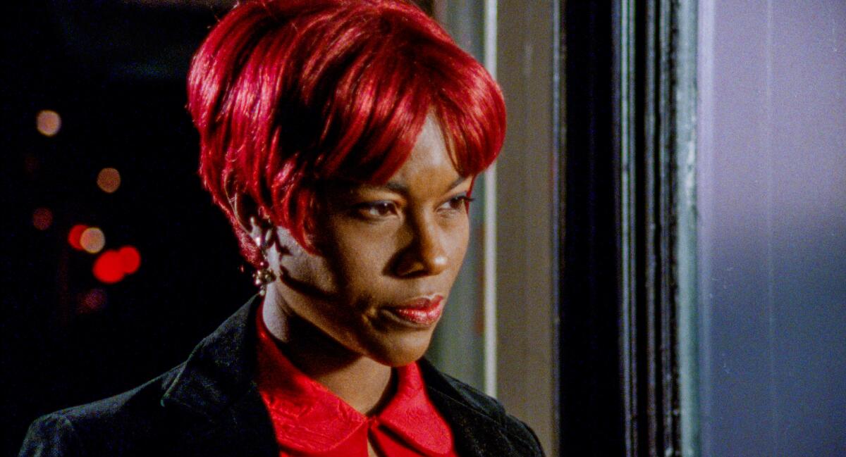 A Black woman in a short red wig stands in a doorway looking intently ahead
