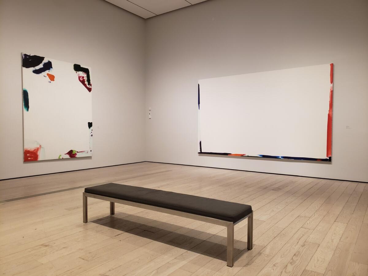Two paintings hang on a wall in a museum, with a bench before them