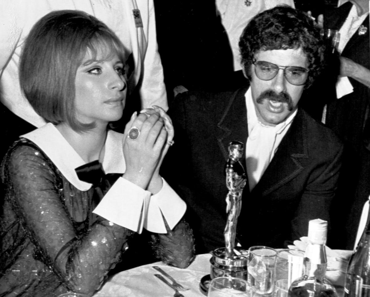 In a black-and-white 1969 photo, Barbara Streisand, left, sits at a table with Elliott Gould with an Oscar in front of her