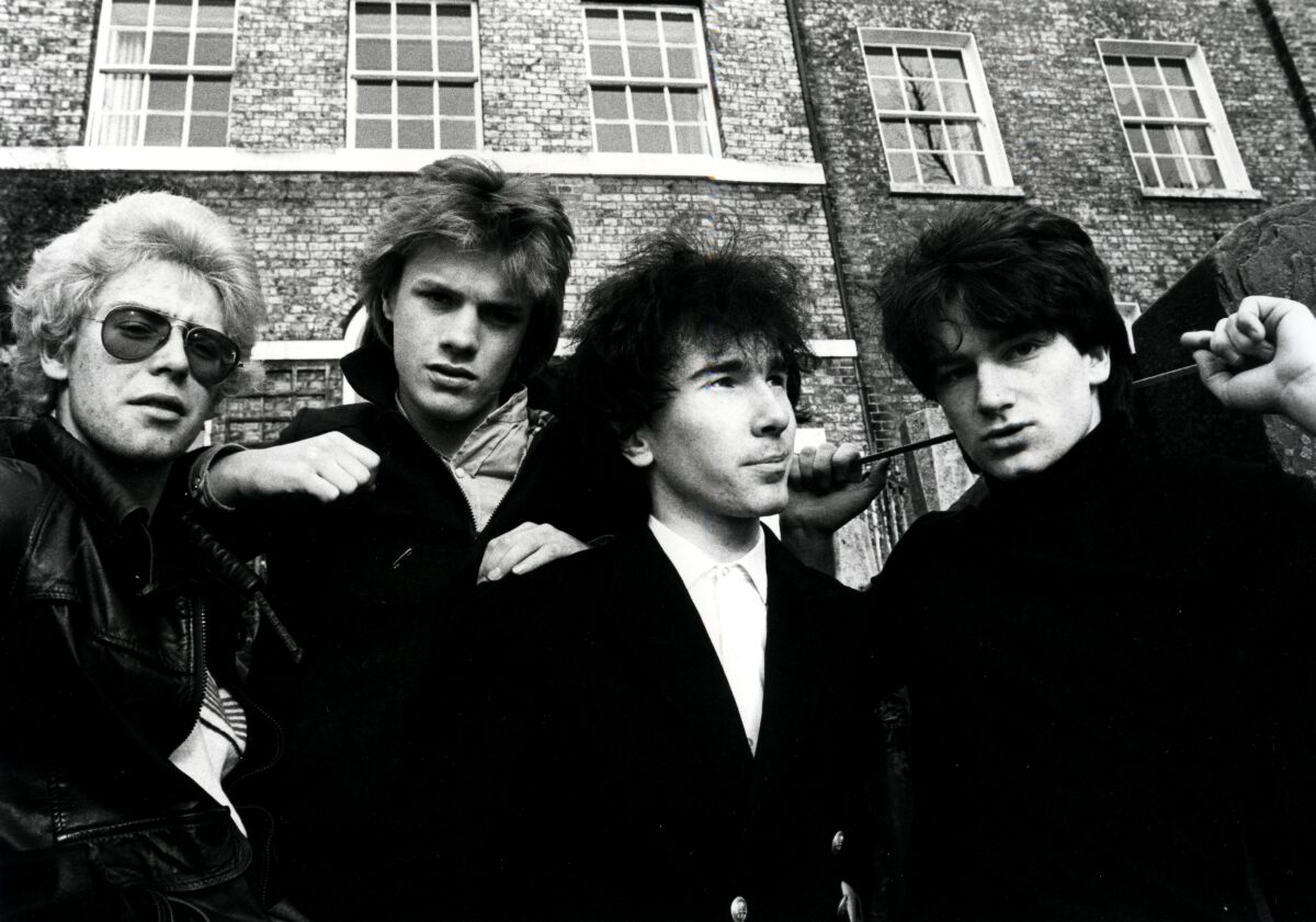 U2 as young rockers with shaggy hair, dressed in dark outerwear in an arty black-and-white outdoor image