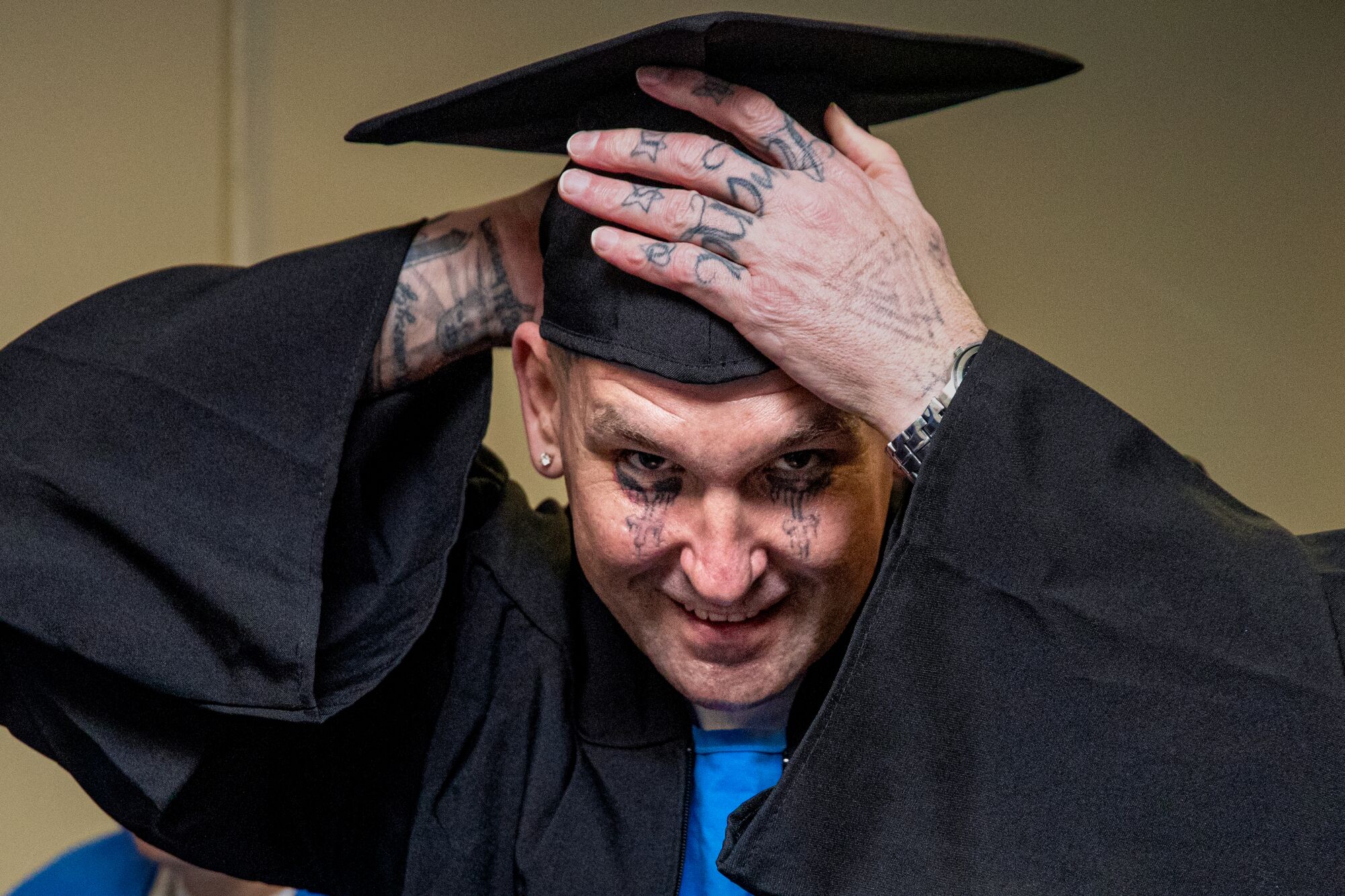 A man with face and hand tattoos looks at the camera while adjusting his black graduation cap. 