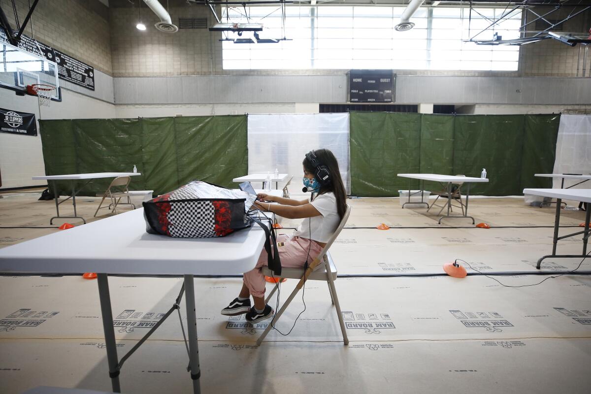 A lone young girl in a mask and a headset works on a computer while sitting at a folding table inside a recreation center