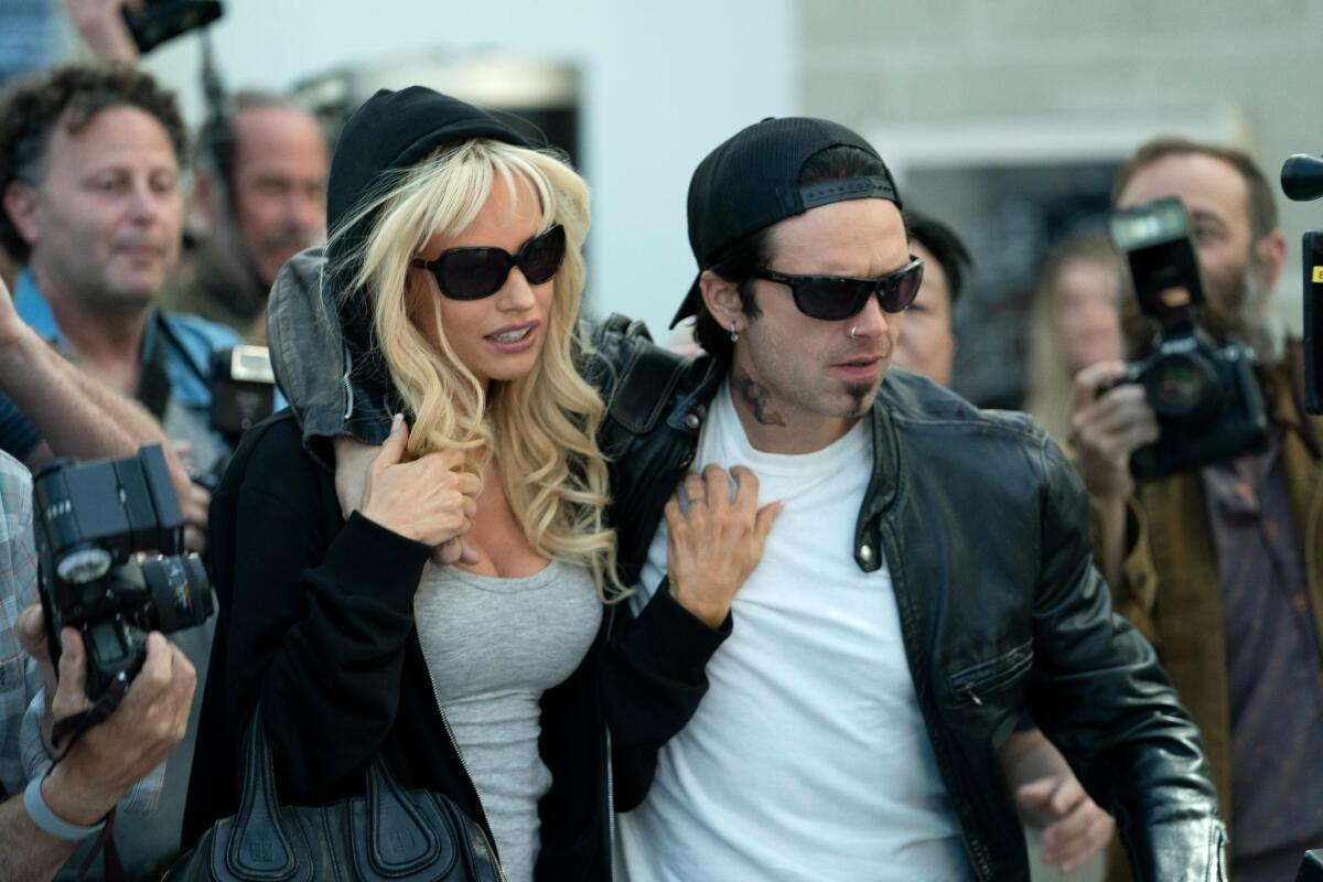 A couple in black leather jackets and sunglasses, with a crowd of photographers behind them.