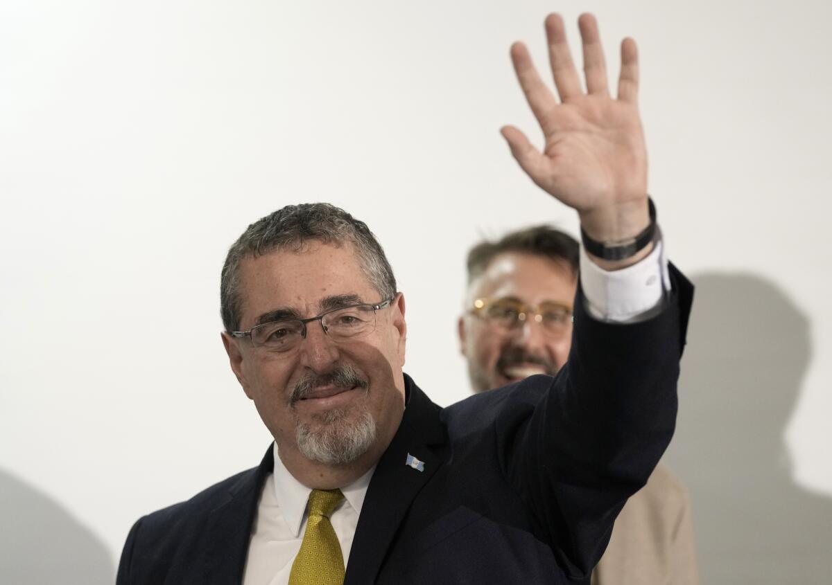 Then-presidential candidate Bernardo Arévalo pictured from the chest up, waving as another man smiles behind him