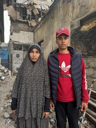 A woman in a gray and black cloak-like robe, her hair covered, and a teenage boy in red and black clothes stand amid rubble