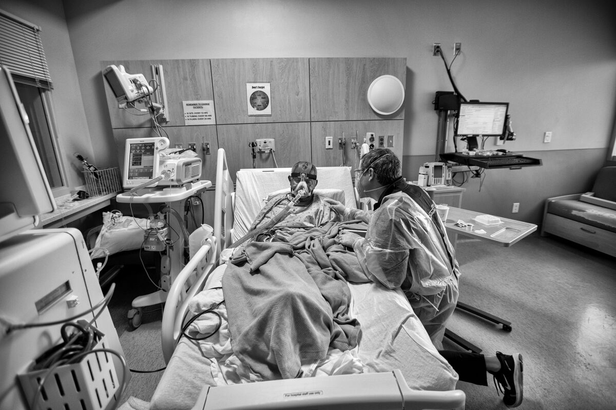 A man in protective gear kneels next to a person in a hospital bed breathing through a respirator.