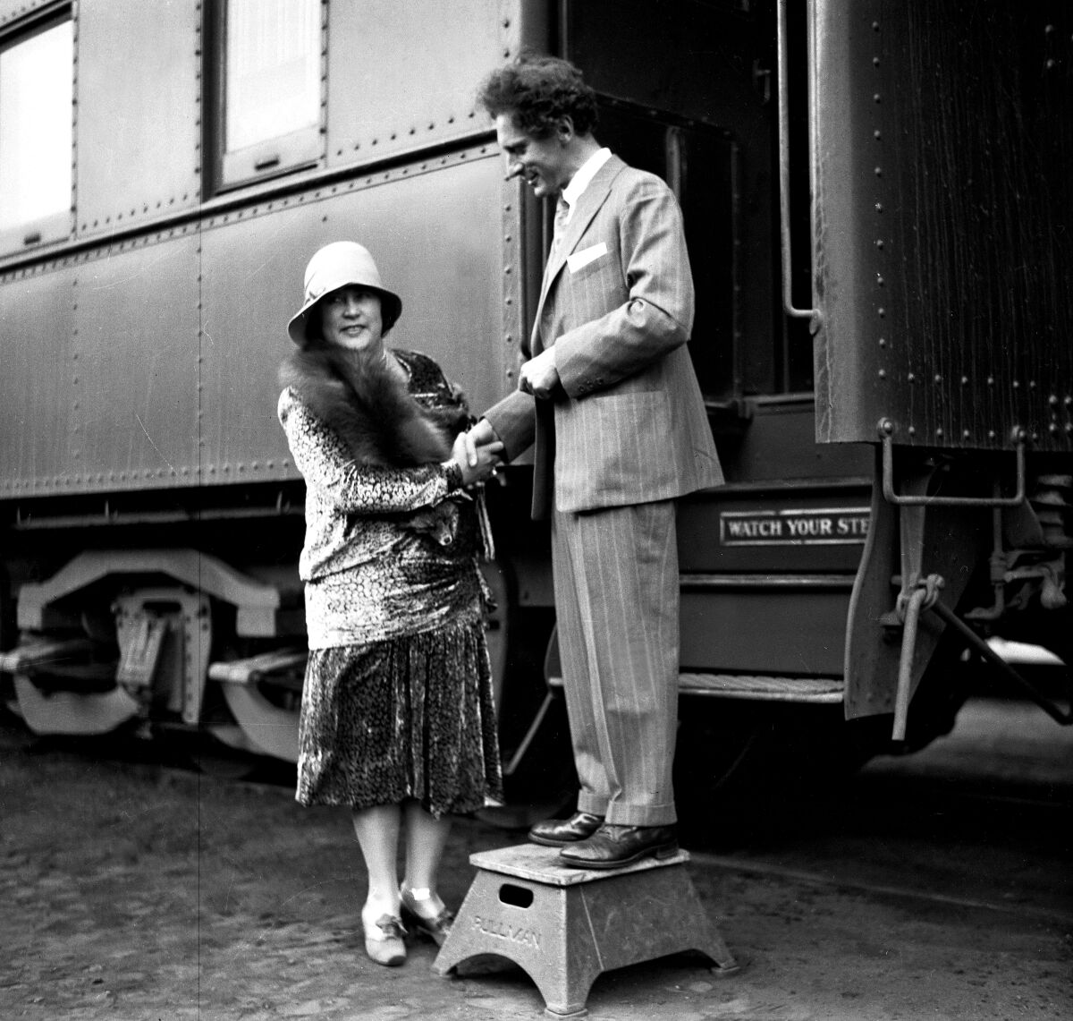 A man descending a train stands on a step stool and holds a woman's hands.