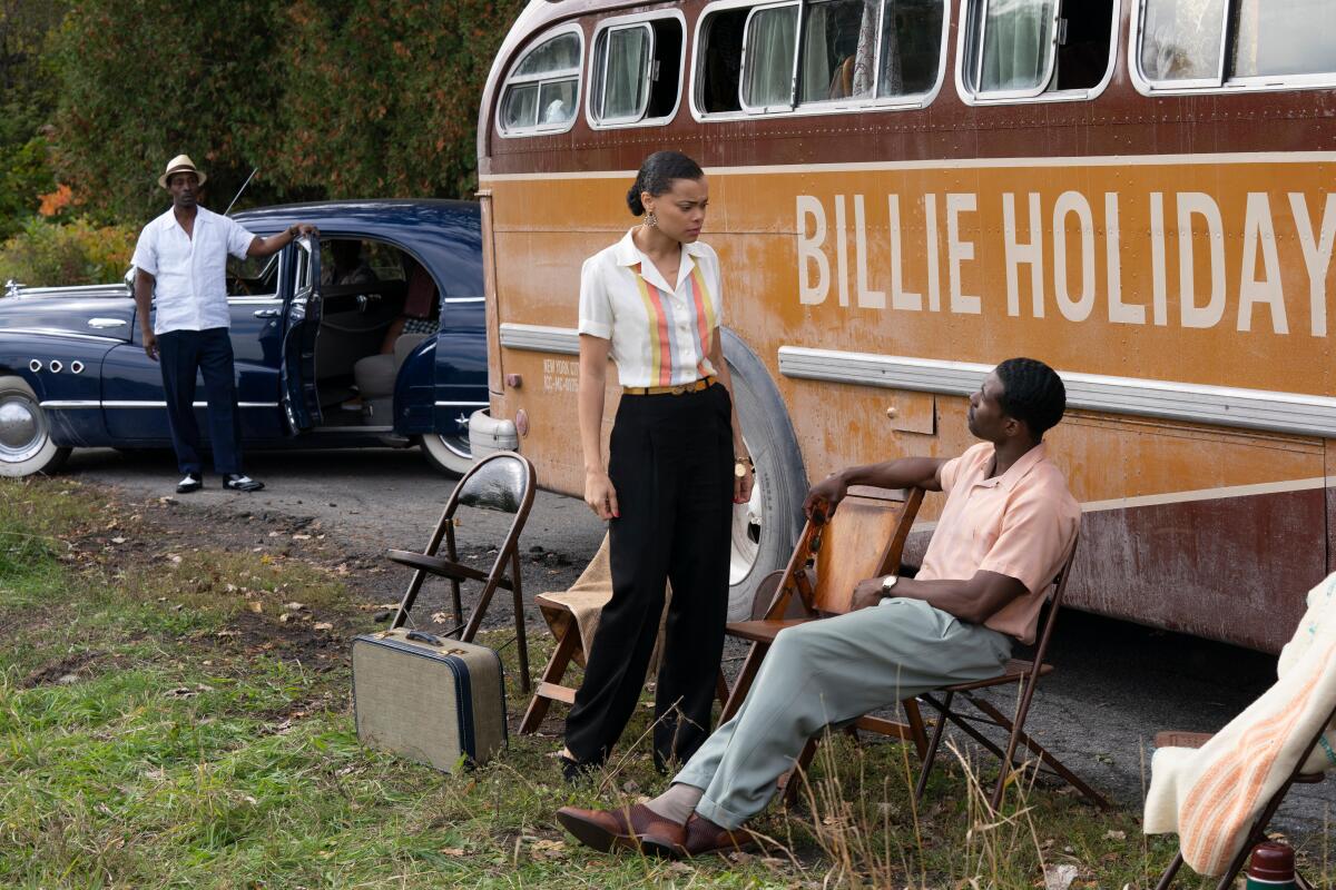 Andra Day as Billie Holiday and Trevante Rhodes as Jimmy Fletcher in a movie scene in the grass  near a parked bus.