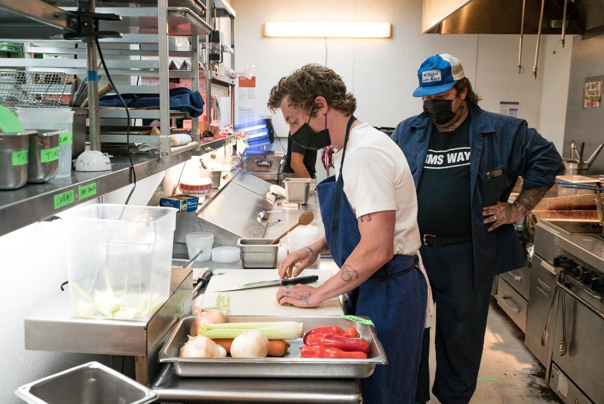 Matty Matheson observes actor Jeremy Allen White at a cutting board in a restaurant kitchen.