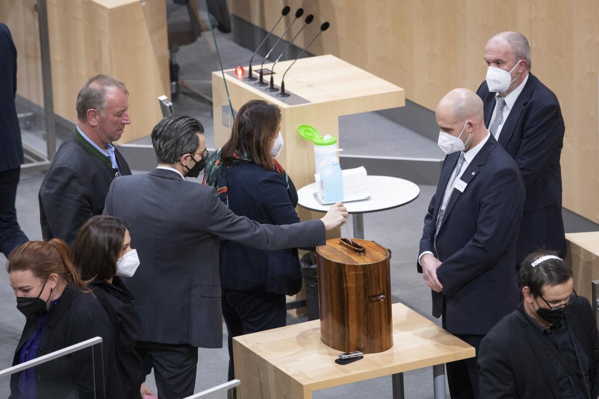 A man in a suit and face mask puts a card in a wooden box on a table with others around him