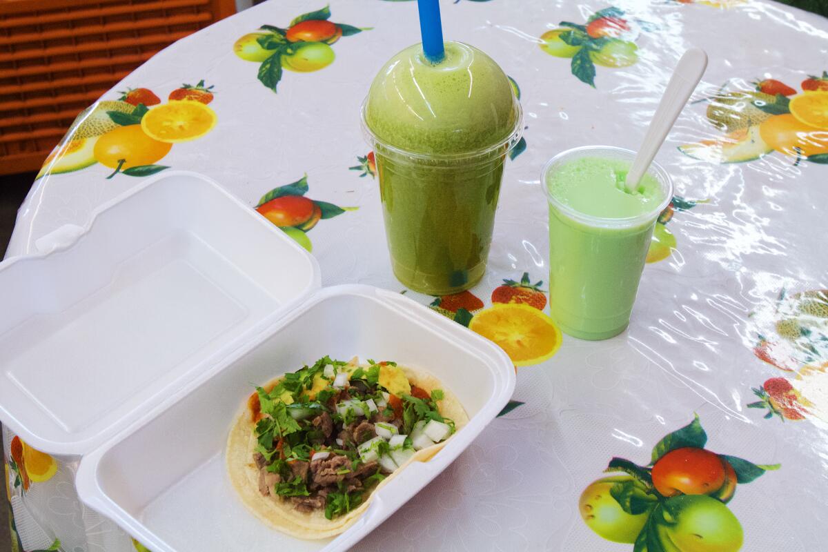 A green juice, a cup of pistachio-flavored Oaxacan Jello, and a tasajo taco on a table with a fruit-patterned tablecloth