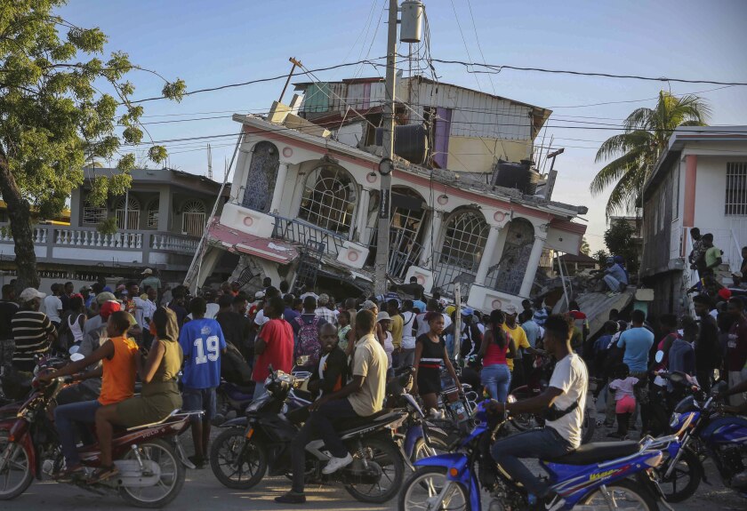 People bike and walk by a toppled two-story building in Haiti.
