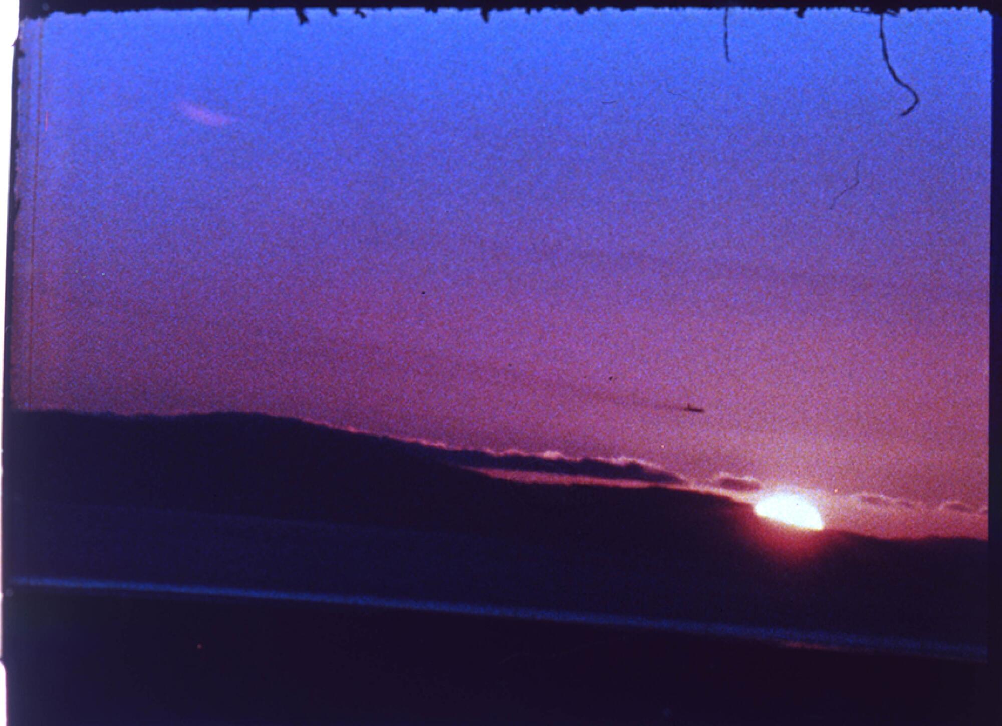 A still of a 16mm film shows the sun setting in fiery purple radiance behind some clouds