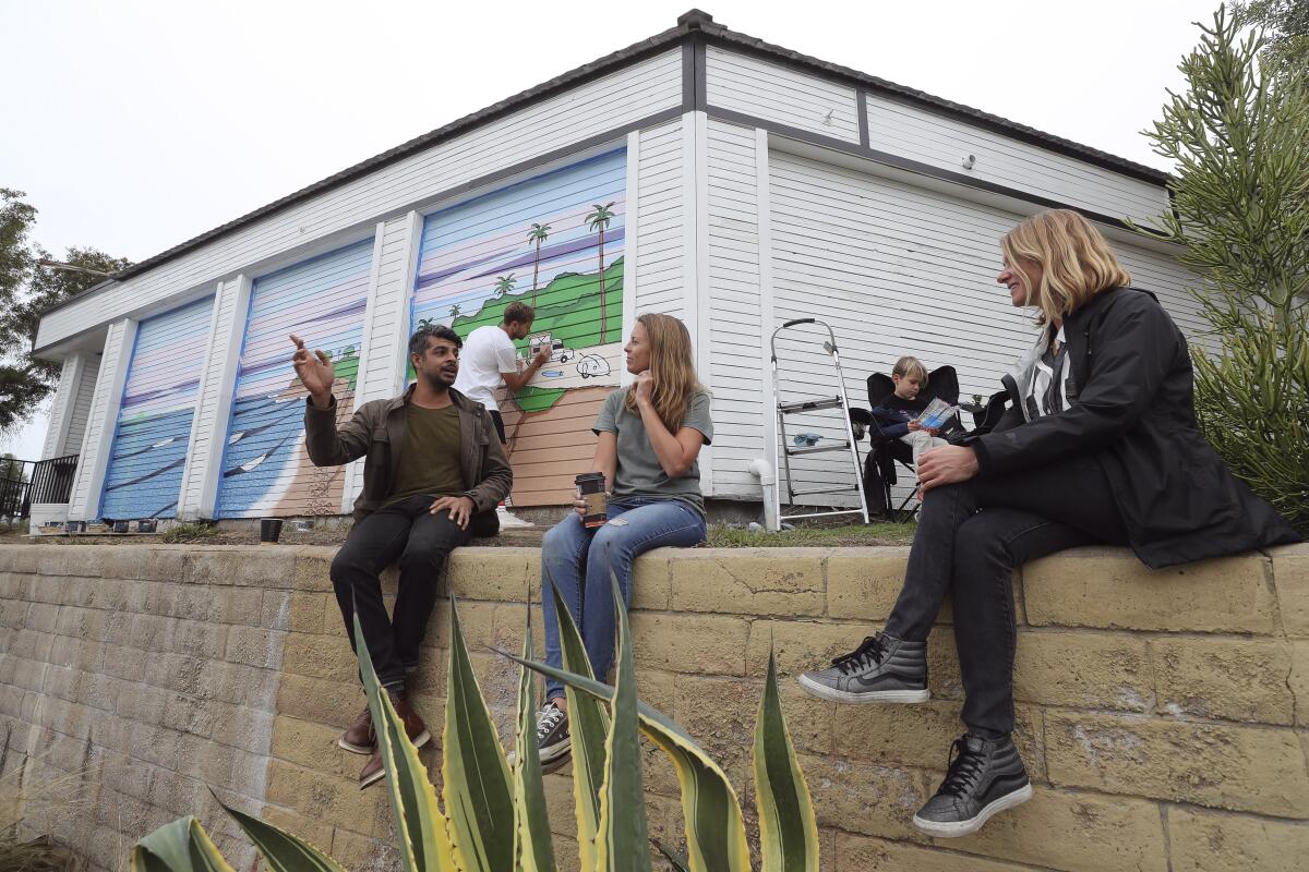 Lora Bodmer-MacDowell, right, her son Wilson MacDowell, 6, artist Steph Blank, and owner of the Cardiff 7-Eleven convenience store Harbir Virk, left, sit at the back of the 7-Eleven as Swedish artist Jonas Claesson, background, works on a mural-painting inspired by a watercolor, titled "More Green Flashes", that was done by Bodmer-MacDowell's husband Tate MacDowell, 39, a Cardiff resident who died of cancer in October, on Tuesday, December 3, 2019 in Encinitas, California.