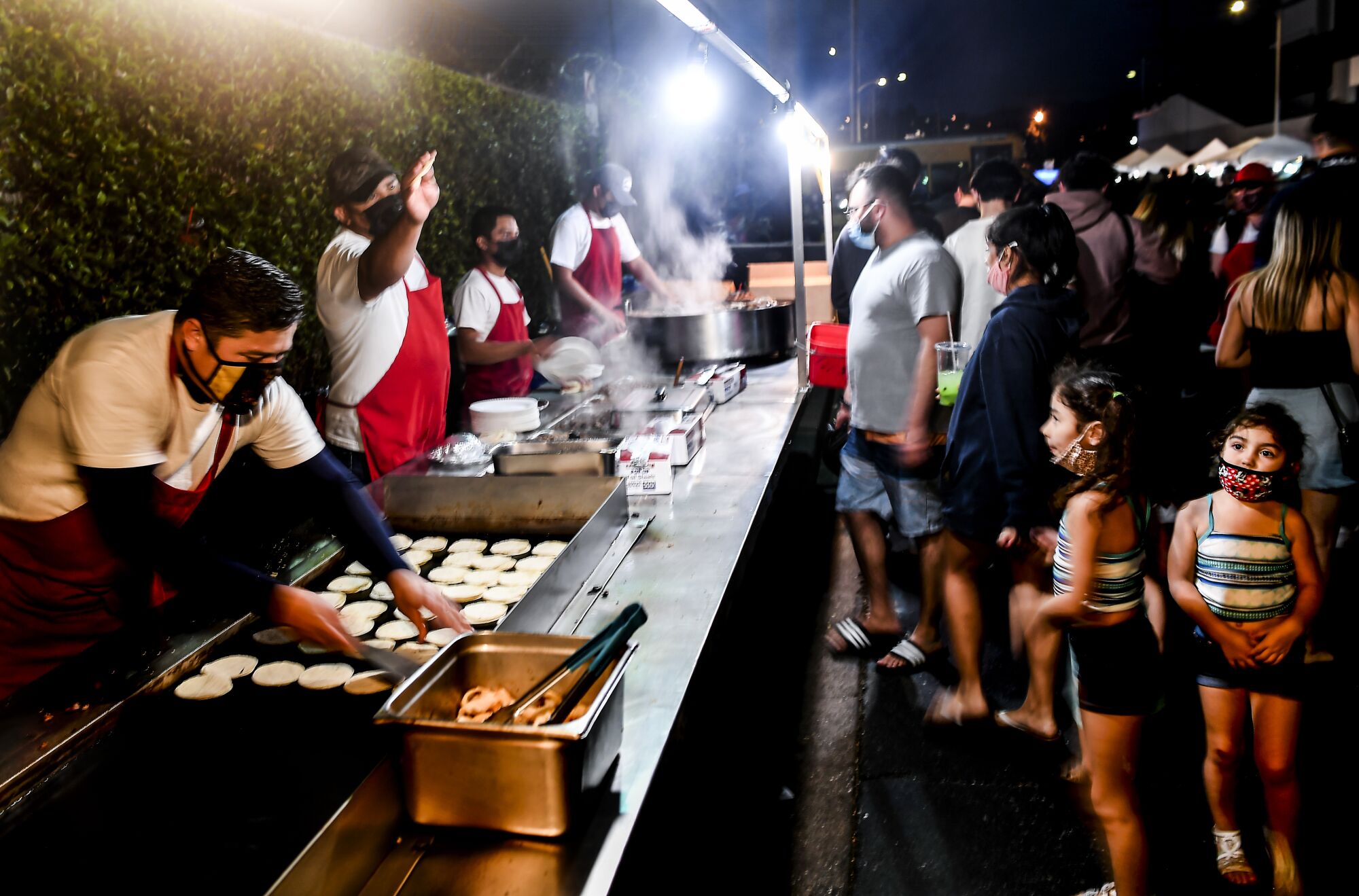 Adults and children wait to be served at an outdoor taco stand at night.
