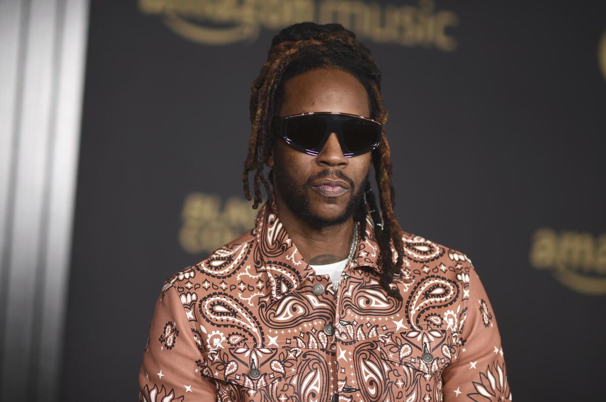 2 Chainz wears a brown, black and white patterned shirt with black sunglasses as he poses for photos at a red carpet event.