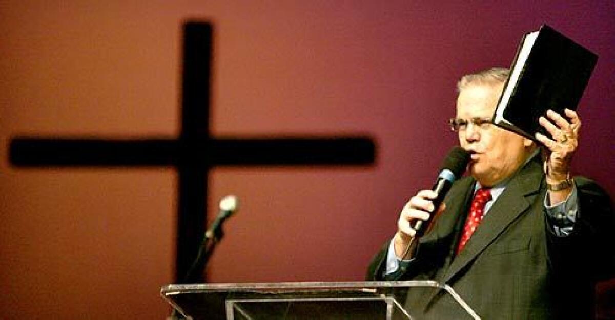 Evangelist John C. Hagee speaks while holding a book and there is a cross near him.