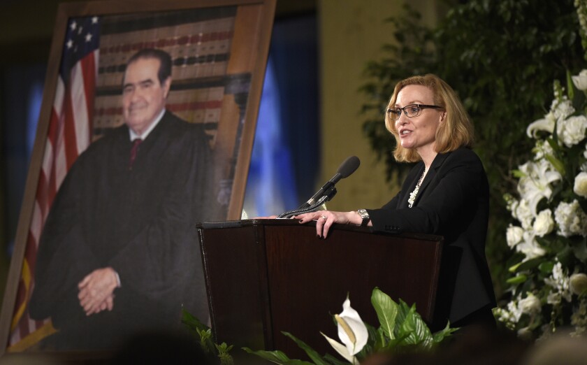 A woman speaks at a lectern before a large portrait of Antonin Scalia 