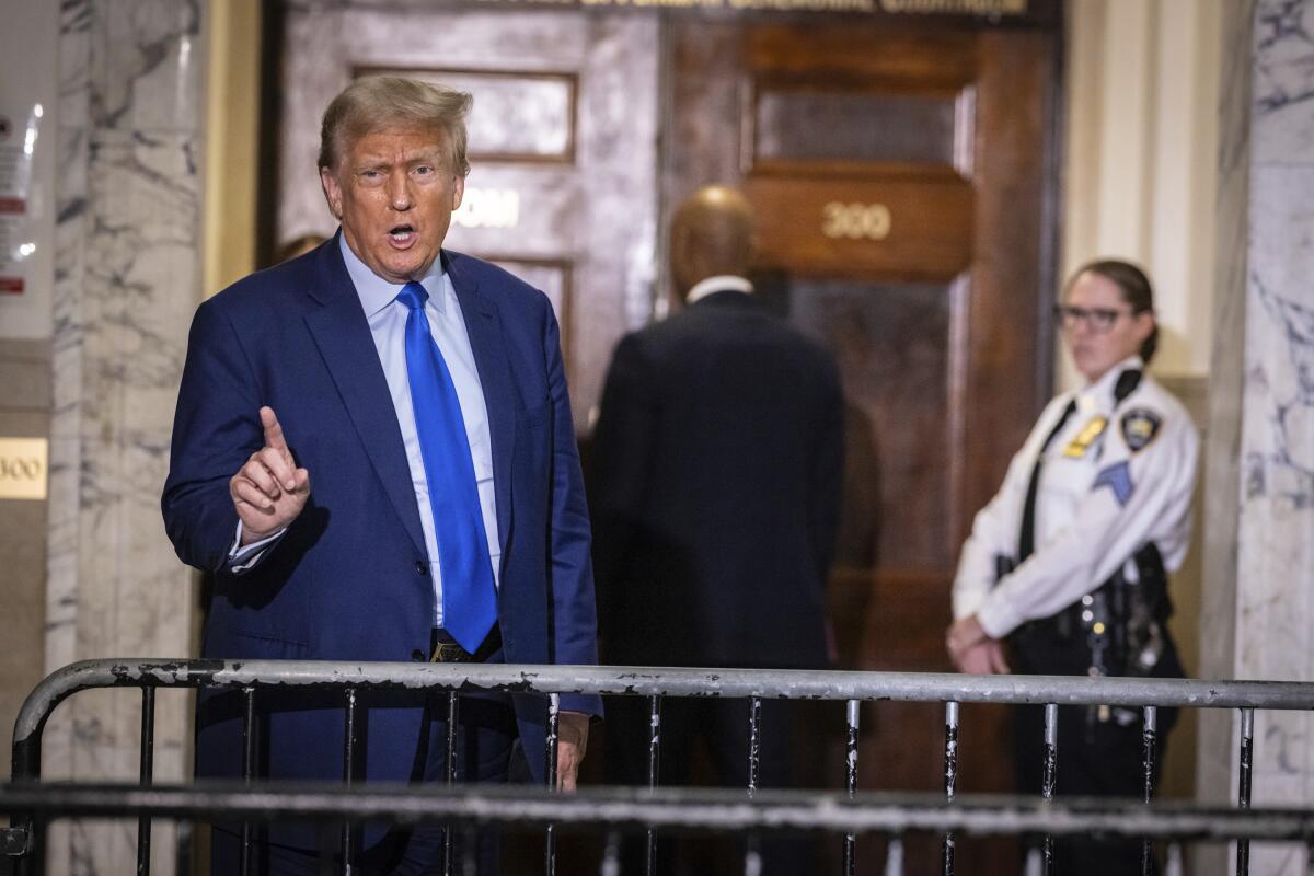 Ex-President Trump raises a finger as he speaks from behind a barricade outside large wood doors as a security guard looks on