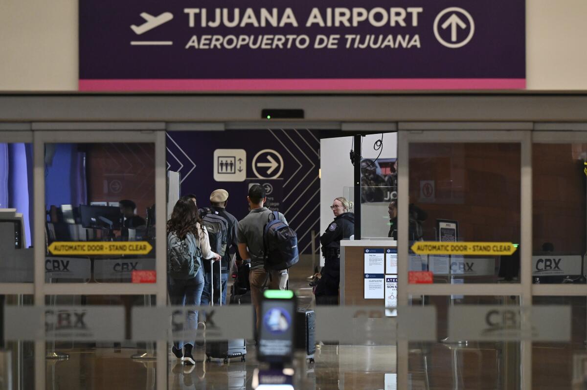 Travelers with their luggage inside a building, with a sign above the doors that says Tijuana Airport