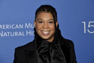 Punkie Johnson with braids wearing a black satin shirt and black suit jacket against a blue backdrop