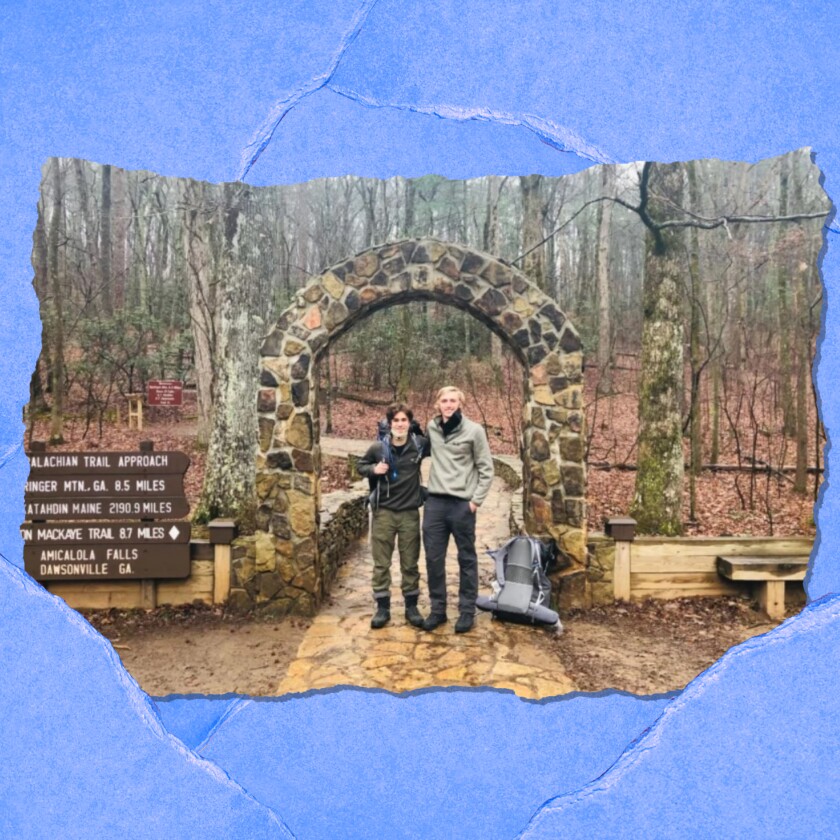 Two young men stand under a stone archway in a forest next to a wooden sign.
