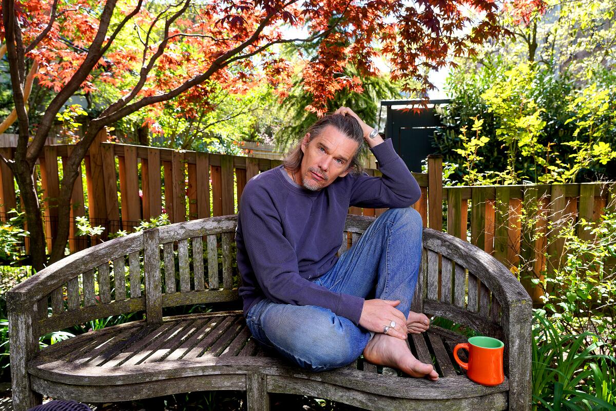 Actor Ethan Hawke on a wooden bench with trees behind him in Brooklyn. 
