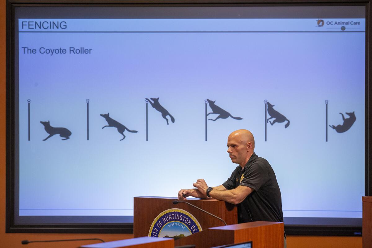 A man speaks at a lectern in front of graphics showing a coyote being blocked from jumping over a wall