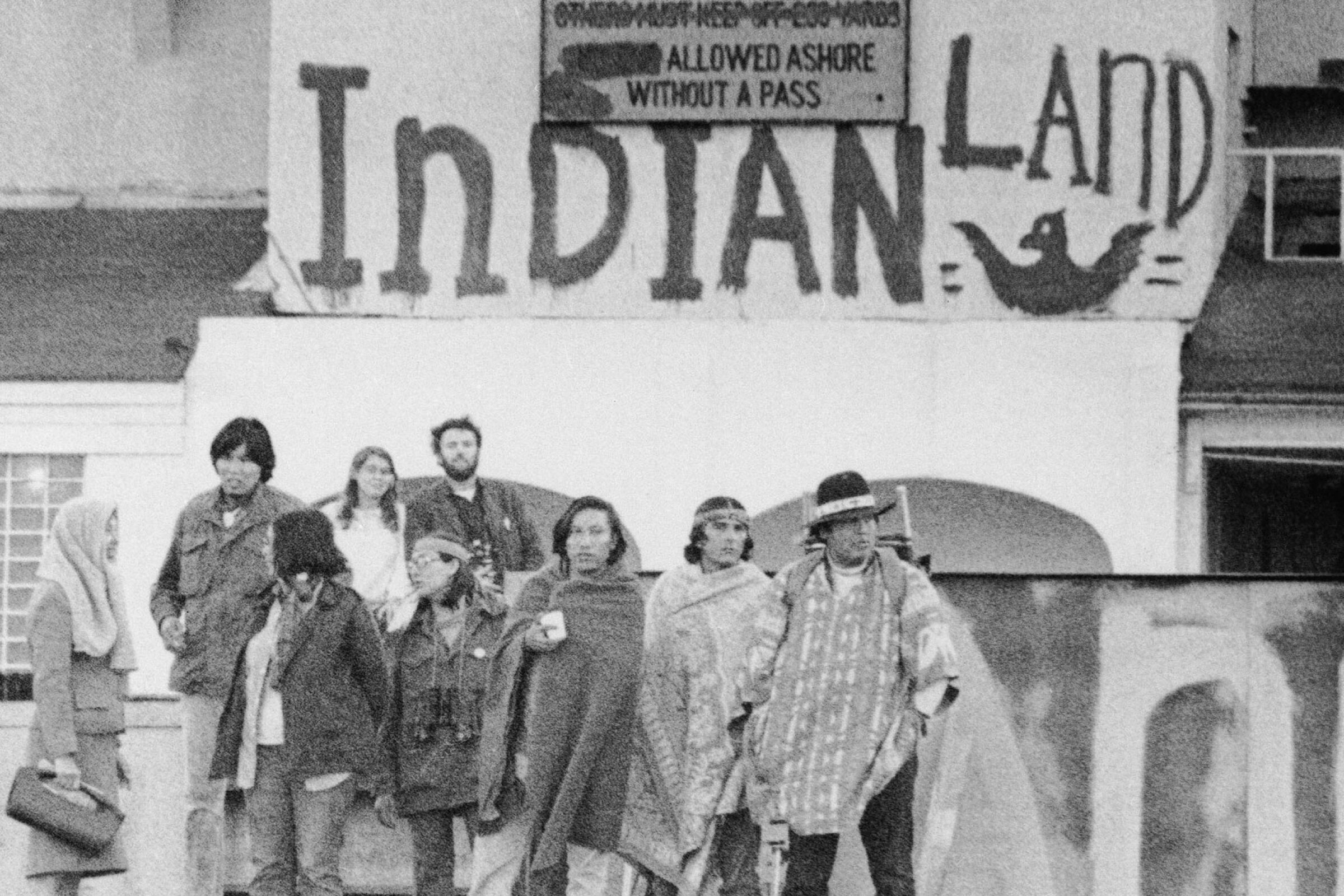 A group of Indigenous people stand on the dock at Alcatraz under signs that read "Indians Welcome" and "Indian Land"