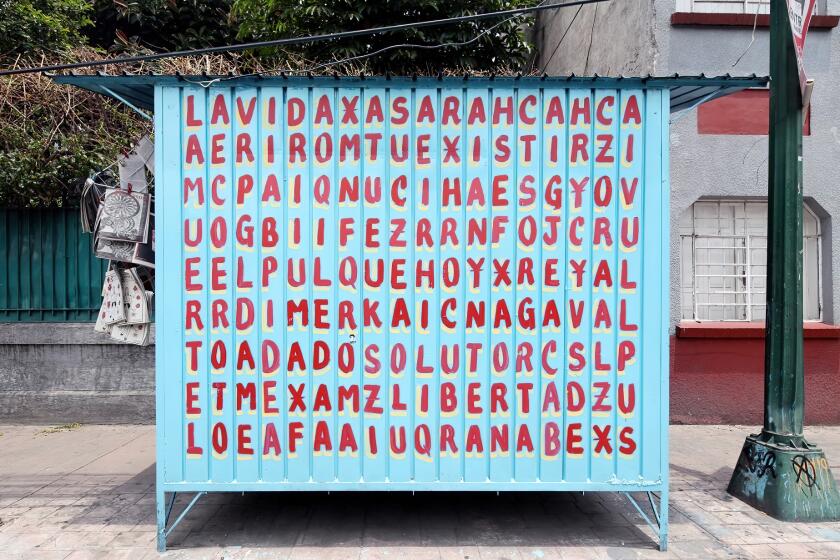 A blue magazine kiosk is covered in a grid of hand-painted letters that reveals phrases like "La Vida" and "El Pulque"