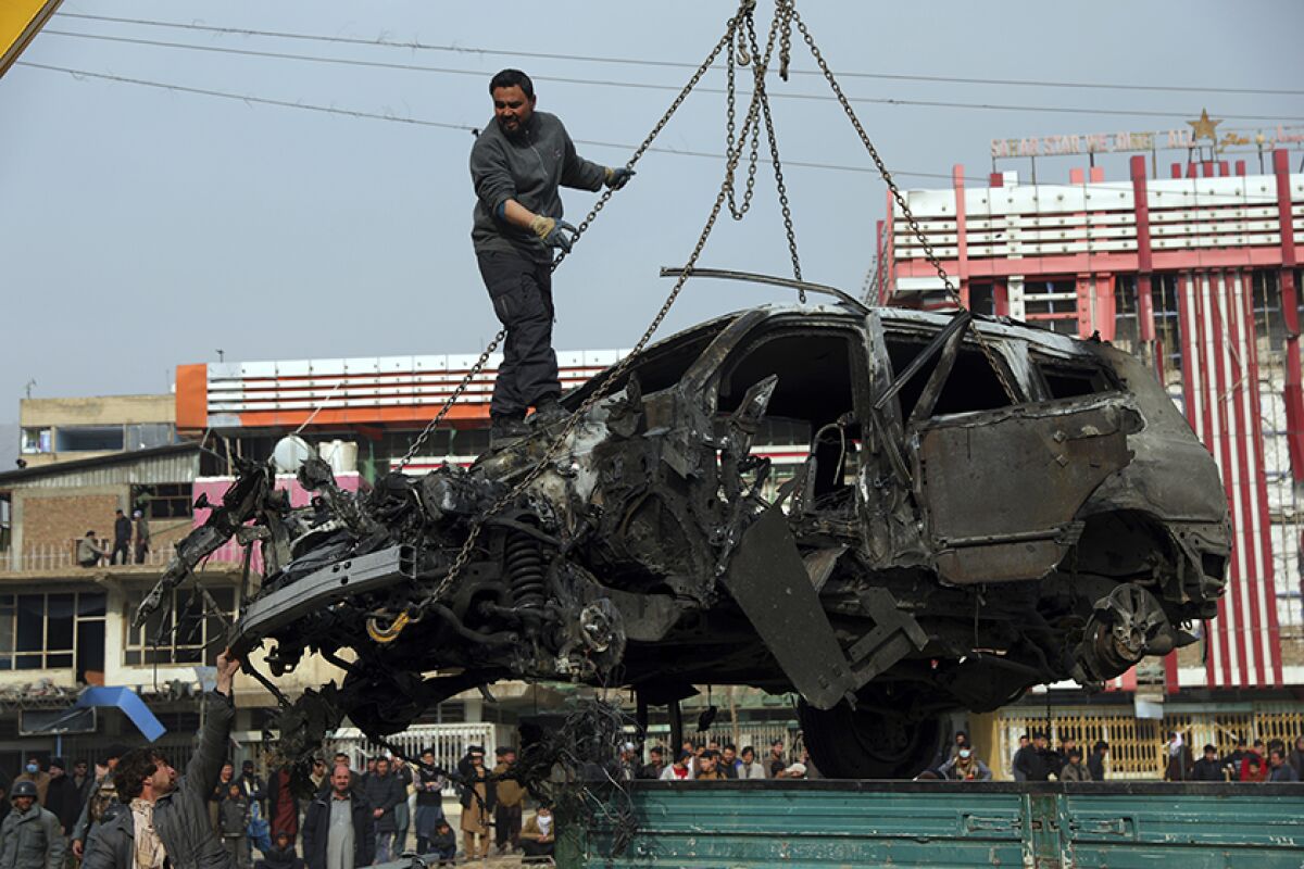 A man stands atop a burned and destroyed car that's being lifted by chains from the ground.