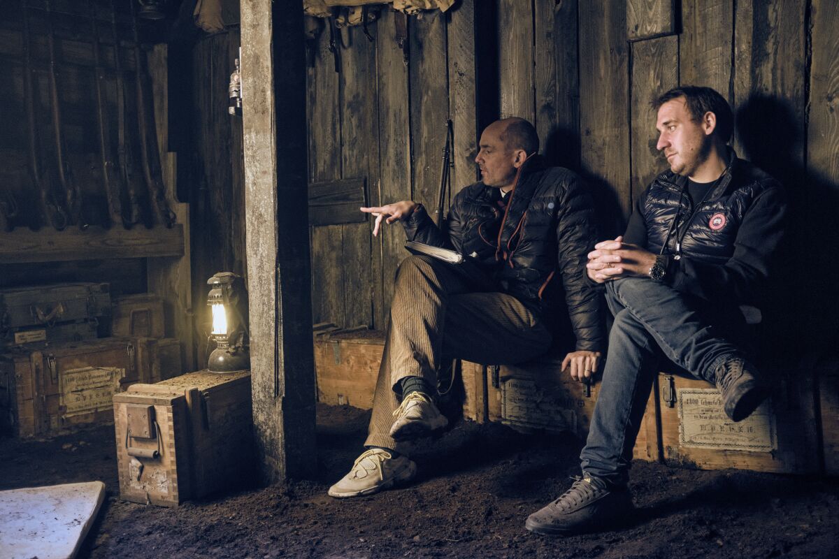 Two men sit and talk in a rustic set on the film "All Quiet on the Western Front."